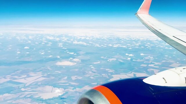 Airplane window view above clouds and agricultural fields with visible wing and engine during flight. Scenic aerial landscape from passenger seat in clear sky. 4K 60fps.