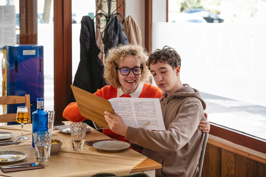 Middle aged mother and teenage son reading restaurant menu together and smiling while choosing food at table