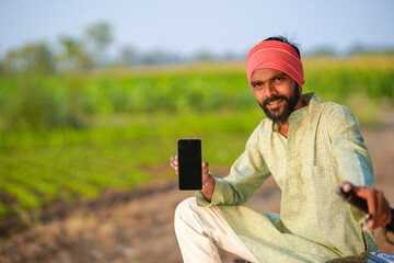 Fototapeta premium young indian farmer showing smartphone screen sitting at green agricultural field