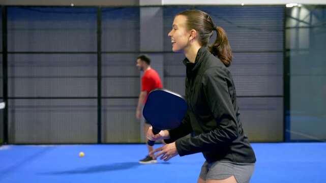 Focused young woman playing a pickleball match with her partner on a blue indoor court. The athletic female player hits the yellow ball with her paddle during an intense and competitive game
