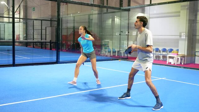Young athletic woman hitting a forehand shot during a friendly pickleball match with her male partner on a modern blue indoor court, showcasing teamwork and an active, healthy lifestyle