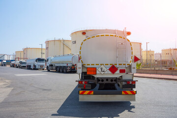 Several white tanker trucks are parked in a line on an asphalt road at a large industrial chemical...