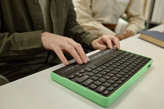 Caucasian young adult man using braille keyboard while sitting beside middle aged Caucasian woman in library setting, hands actively reading and typing on assistive device