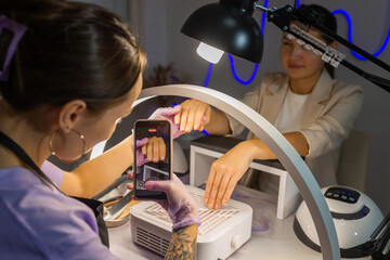 Nail technician photographing finished manicure on female hands using smartphone under ring lamp in beauty salon. Professional nail care, content creation, social media and manicure service concept.
