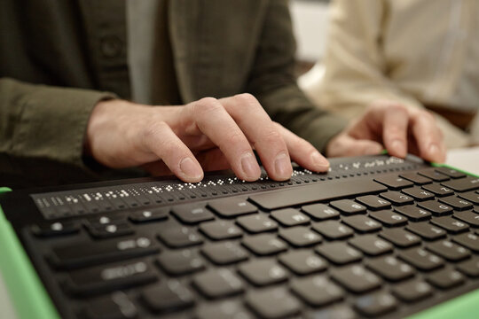 Caucasian young adult man reading braille display with both hands in library setting, focusing on tactile text while sitting at desk, another partially visible in background