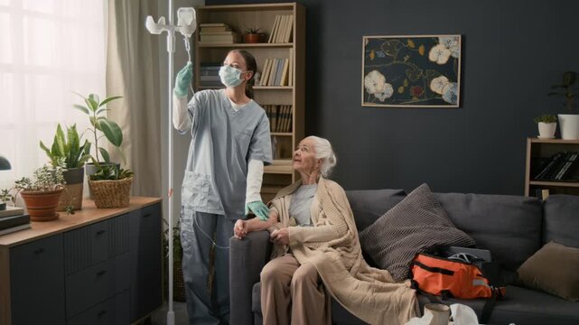 Young female physician wearing grey scrubs inserting IV into arm of sick elderly woman sitting on couch while providing home care services