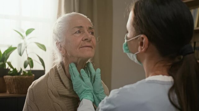 Over the shoulder rack focus shot of brunette young nurse wearing rubber gloves palpating lymph nodes of senior ladys neck during home medical examination
