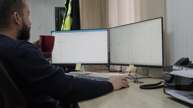 Side view of man working at desk with dual monitor computer setup in modern office environment. He drinks coffee while looking at two large screens displaying text documents
