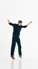Young man in black sweater and jeans captured mid jump with arms raised on white background. Concept of freedom, youth energy, casual fashion, self expression and modern lifestyle.