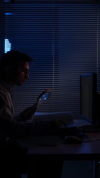 Vertical video man in silhouette uses a magnifying glass to examine a computer screen in a dim office, evoking cyber investigation, digital forensics, data analysis and online security