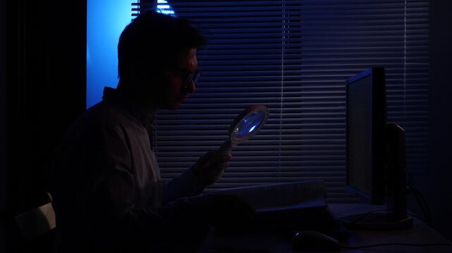 Man in silhouette working an investigation, meticulously examining evidence with a magnifying glass while sitting in front of a computer screen in a dark office at night
