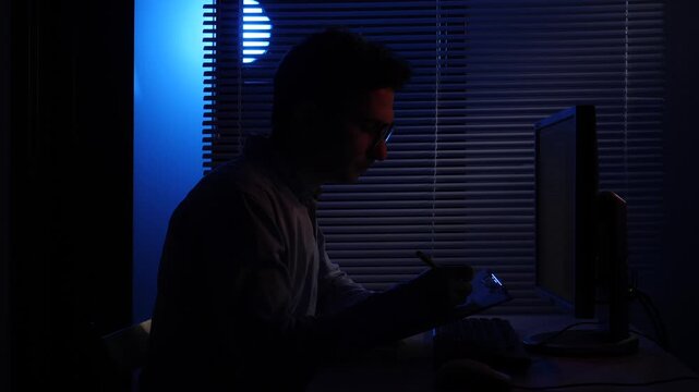 Young man wearing optical glasses sitting at a desk, writing notes in a notebook next to a computer monitor, illuminated by blue light from a window with blinds at night