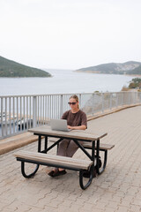 Fototapeta premium Digital nomad working on laptop by the sea, portable solar panel on backpack, remote work lifestyle, sustainable travel, coastal scenery, work from anywhere concept, copy space.