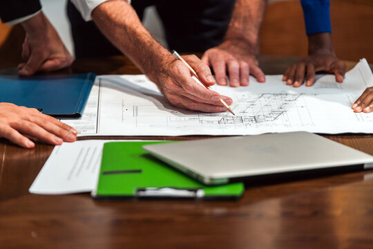 Team collaborating over architectural blueprints on a table, marking plans with a pencil beside a laptop and folders during a project meeting.