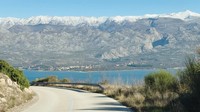 Handheld camera view from a moving vehicle, a curvy coastal road above the blue Adriatic Sea with the massive Velebit mountain range covered in snow. Bright winter sun and scenic Croatian coastline