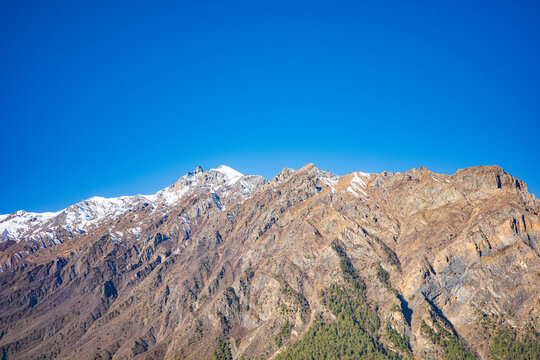 Wide panoramic view of a rugged alpine mountain range with scattered snow patches and steep rocky ridges beneath a vivid blue sky. The dramatic high-altitude terrain and pristine wilderness scenery