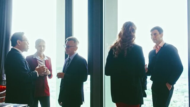 Professionals in suits mingle and chat during a corporate networking reception by large windows with a city view, holding drinks and engaging in conversation.