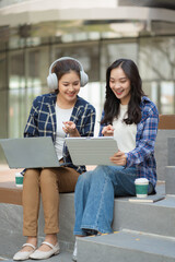 Naklejka premium Two young Asian female college students studying together outdoors in a park, using laptop, tablet, and notebooks while preparing for exams or working on a group project.