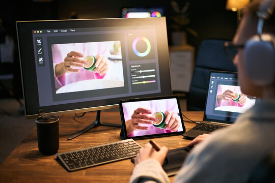 Caucasian young adult man using graphic tablet and stylus editing product photo on computer and tablet, wearing headphones, focusing on digital retouching process at desk