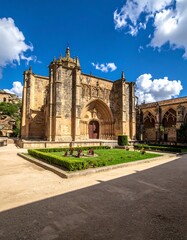 A historic stone building with a large archway and a well-manicured garden under a blue sky with white clouds