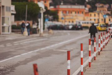 A bicyclist pedals down a quaint road lined with red and white poles, surrounded by colorful buildings in a charming coastal town, basking in the warm afternoon light