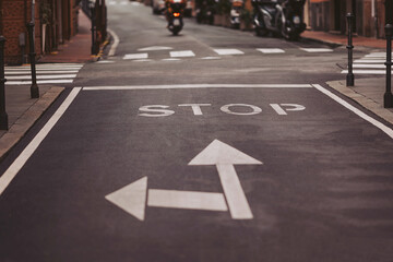 A stop sign painted on the asphalt directs vehicles at a lively crossroads. Motorcycles pass by, while street lamps and colorful buildings create an inviting city scene