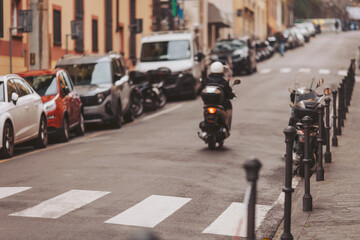 A busy street showcases a scooter moving alongside rows of parked cars under a clear blue sky. Paved sidewalks and zebra crossings add to the lively scene