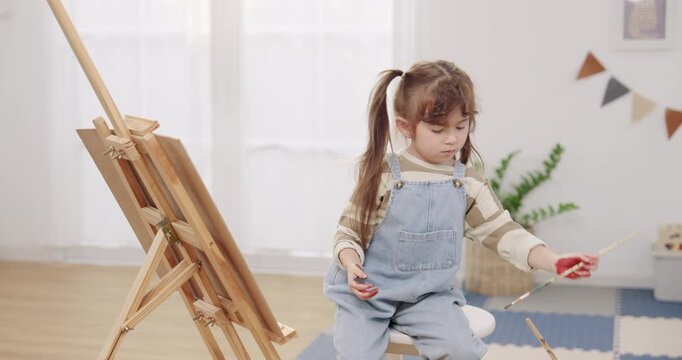 Asian preschooler daughter pauses painting, raises paint covered fingers near easel canvas, playtime in living room, kids art therapy and expression, fine motor skill practice, weekend learning