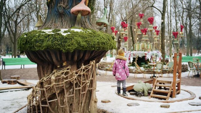 Winter park scene with children playing near a whimsical treehouse style structure decorated with moss and fairy tale elements