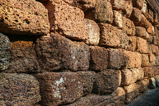 Perspective view of ancient laterite stone wall with deep shadows enhancing texture and depth. Weathered red brown blocks create dramatic historical background atmosphere.