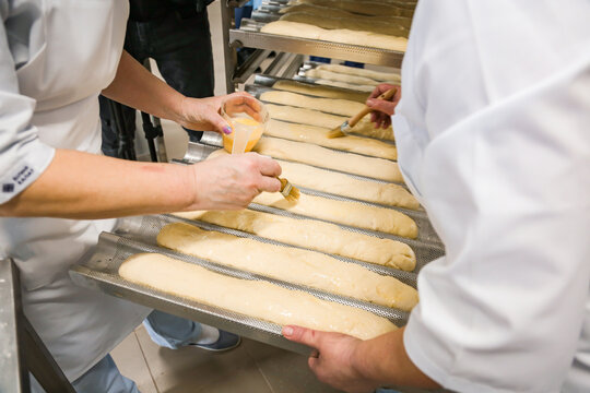 Women brushing baguettes with egg wash in a bakery kitchen before baking. Traditional bread preparation process in professional food production environment.