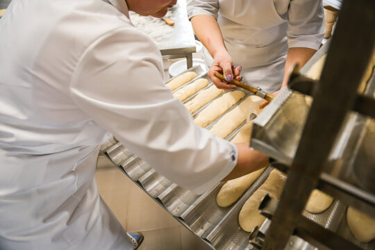 Women brushing baguettes with egg wash in a bakery kitchen before baking. Traditional bread preparation process in professional food production environment.