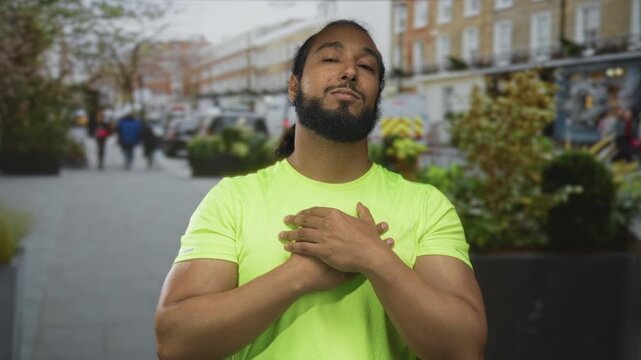 Man with hands on chest on street wearing neon green shirt, arms slightly open toward camera while pedestrians pass behind him; gratitude sincerity.