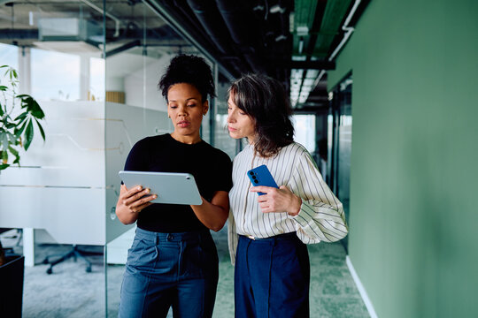 Two diverse businesswomen collaborating and sharing ideas with a digital tablet while walking in a modern corporate office corridor