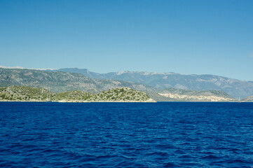 Fototapeta premium A wide horizontal landscape of the Turkish coast near Kekova. Deep blue sea waves in the foreground contrast with rocky islets and massive mountain ranges under a clear sky.