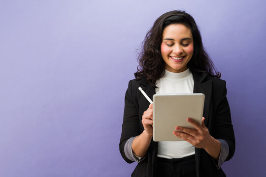 Latin businesswoman smiling, using a tablet with a stylus, creating or editing content against a vibrant purple background