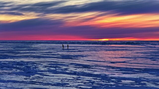 People ice skating on frozen sea at vibrant sunset with colorful sky, Distant people silhouettes ice skating in slow motion aerial view