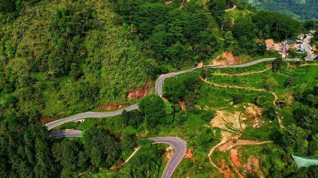 Aerial view of a winding mountain road on a lush green hillside near Ambuluwawa Tower in Sri Lanka. The drone shot captures switchback curves and surrounding vegetation from above, showing the steep s