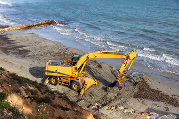 Obraz premium A yellow excavator operates on a sandy shore near the Mediterranean sea in Spain, engaged in earthmoving work near the shoreline.