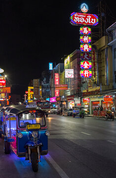 Bangkok, Thailand - 02 02 2026: Vertical night shot of a street in Bangkok's Chinatown. In the foreground, a blue and red tuk-tuk is parked, featuring bright blue neon underglow lights.