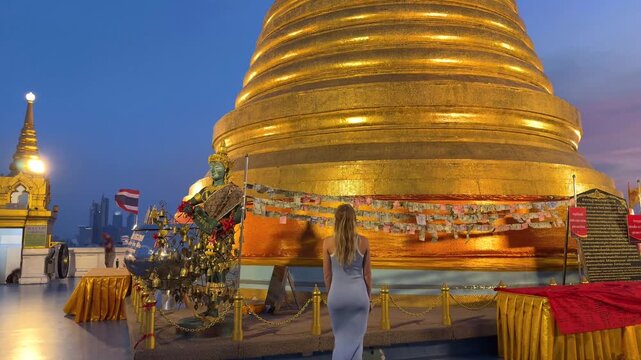 Woman walking toward the Golden Mount temple (Wat Saket) in Bangkok, Thailand at sunset. Iconic golden stupa and spiritual landmark reflecting Thai culture and travel experience.