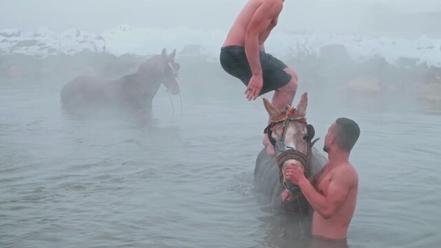 A man performs a backflip into a steaming natural hot spring while others bathe horses in winter. Snowy landscape, rising mist and dramatic action create a unique outdoor scene.