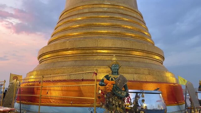 Golden Mount temple (Wat Saket) illuminated at sunset in Bangkok, Thailand. Iconic golden stupa and historic Buddhist landmark against dramatic evening sky.