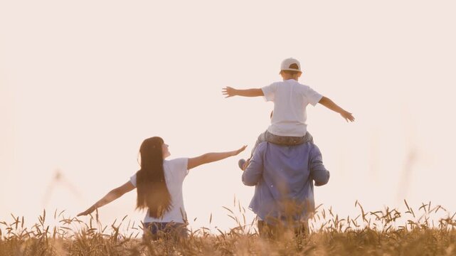 Happy family walking on dry wheat field flying with open hands playing together back view. Cheerful mother father and son fooling relaxing enjoy freedom at sunny sunset harvest natural landscape