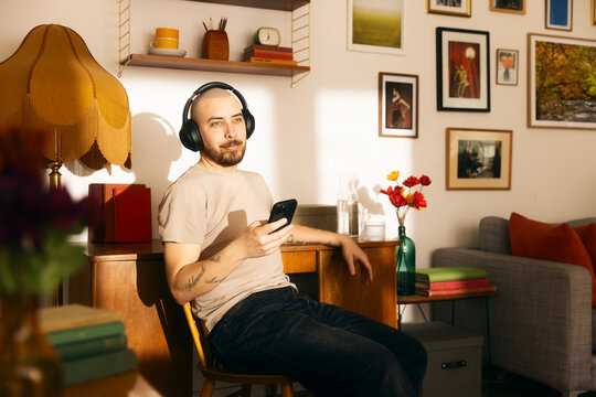 A relaxed man with a shaved head and tattoos sits in a sunlit, vintage-style living room, wearing over-ear headphones and looking at his smartphone while enjoying music or a podcast.