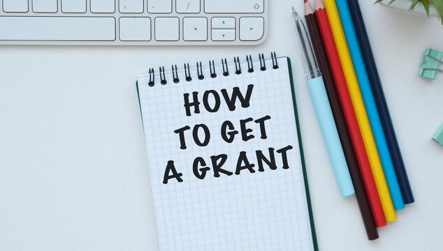 HOW TO GET A GRANT text written in a spiral notebook on a white office desk with colorful pencils.