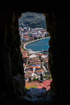 An ancient fortress arrow slit opening frames the vibrant town of Ohrid with its red roofs, extending to the shimmering blue Lake Ohrid and distant mountains.