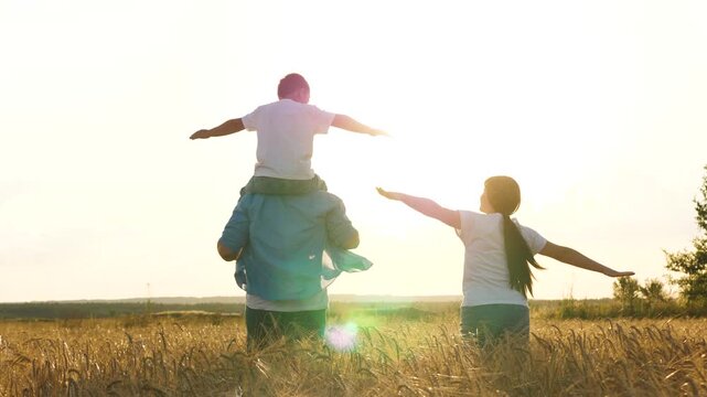 Happy family running at sunset dry wheat field with open hands flying imagination back view. Cheerful mother father and son fooling playing together flight fantasy imagine freedom happiness and unity