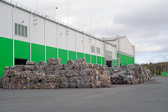 Compressed blocks of plastic bottles and mixed recyclables are neatly piled outdoors in front of a substantial green and white industrial building