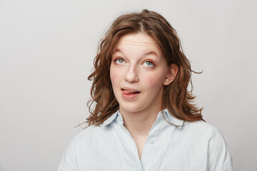 Fototapeta premium Young woman with curly hair and green eyes makes funny face sticking out tongue. Casual white shirt and neutral background create playful and lighthearted mood in studio portrait.